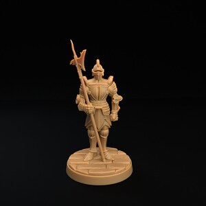 Op de afbeelding: Een miniatuurfiguur van een ridder in volledige bepantsering, die een speer met een scherpe punt vasthoudt. De ridder staat op een ronde basis met een steenachtige textuur.