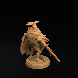Op de afbeelding: Een miniatuurfiguur van een ridder, geschilderd in een lichtbruine kleur. De ridder draagt ​​een harnas en houdt een zwaard en schild vast. De ridder heeft een groot, enkel oog en een kroon van bladeren op zijn hoofd.