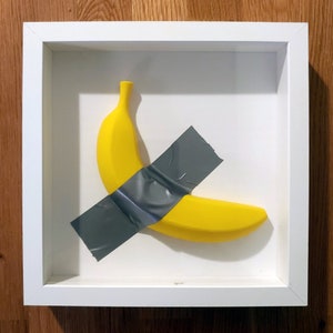 May include: A yellow banana taped to a white background inside a white frame. The banana is taped with grey duct tape.