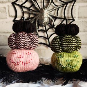 May include: Handmade Halloween-themed knitted pumpkins in pink, green, and black. The pumpkins are stacked and decorated with beaded accents. A black spider web and glittery spider are in the background, with black feathers in the foreground.