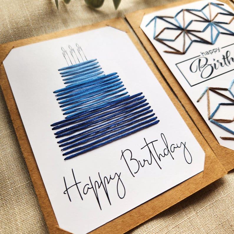 May include: Handmade birthday card with a blue embroidered cake design and three candles. The words "Happy Birthday" are written in cursive. The card is set on a neutral background.
