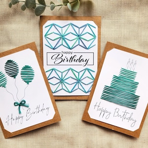 May include: Three handmade birthday cards with white backgrounds and brown borders. Each card features a different teal design: balloons, geometric shapes, and a cake. The words "Happy Birthday" are written on each card.