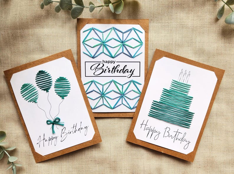 May include: Three handmade birthday cards with white backgrounds and brown borders. Each card features a different teal design: balloons, geometric shapes, and a cake. The words "Happy Birthday" are written on each card.