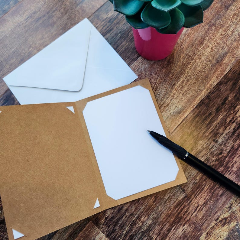 May include: A blank greeting card with a white interior and a brown exterior, alongside a white envelope and a black pen. A small potted succulent in a pink pot is on a wooden surface.