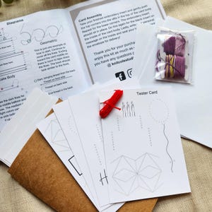 May include: A craft kit with instructions and cards for embroidery. The kit includes a booklet, white cards with geometric designs, red thread, and a small bag of purple and yellow thread. The text on the cards reads "Tester Card".