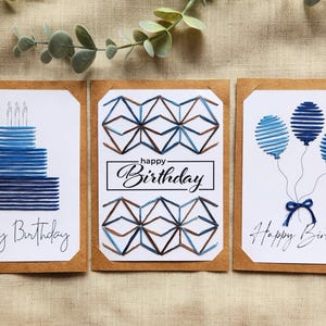 May include: Three birthday cards with watercolour designs. One card features a blue cake with candles, another has geometric shapes, and the third shows blue balloons. Each card has the words "Happy Birthday".