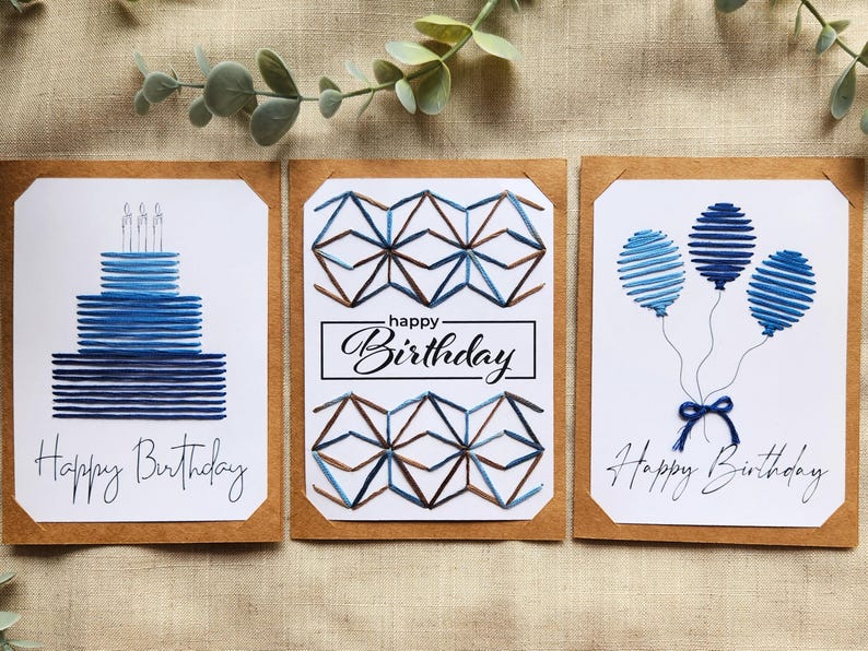 May include: Three birthday cards with watercolour designs. One card features a blue cake with candles, another has geometric shapes, and the third shows blue balloons. Each card has the words "Happy Birthday".
