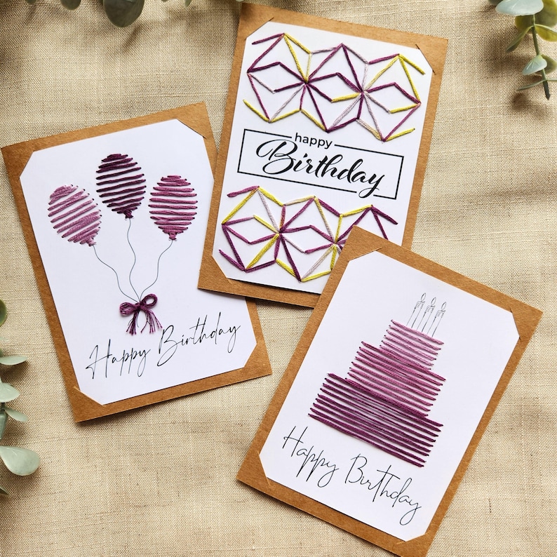 May include: Three handmade birthday cards with white backgrounds and brown borders. The cards feature embroidered designs of balloons, geometric shapes, and a cake. Each card has the words "Happy Birthday" written in cursive.