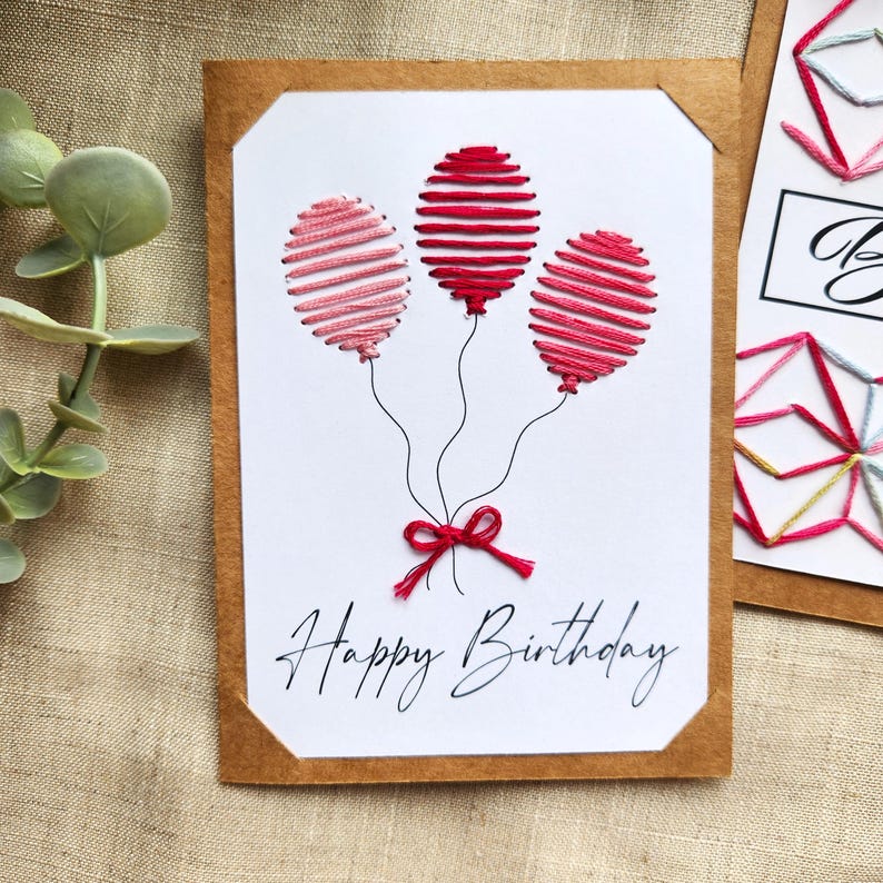 May include: A handcrafted birthday card with three embroidered balloons in red and pink hues. The balloons are connected to a red bow with black threads. The words "Happy Birthday" are written in cursive below the balloons.