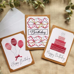 May include: Three handmade birthday cards with brown borders. One card features red balloons, another a cake, and the third a geometric design. Each card has the words "Happy Birthday" in cursive. The cards are on a neutral background.