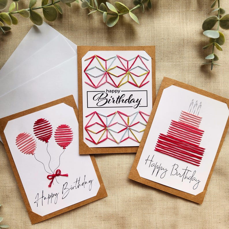 May include: Three handmade birthday cards with brown borders. One card features red balloons, another a cake, and the third a geometric design. Each card has the words "Happy Birthday" in cursive. The cards are on a neutral background.