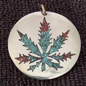 May include: A silver pendant in the shape of a marijuana leaf, inlaid with turquoise and red stones.