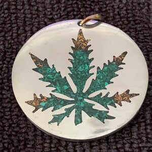 May include: A silver pendant with a green and gold marijuana leaf design.