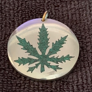May include: A silver pendant with a green marijuana leaf design. The leaf is made of a textured material, possibly turquoise.
