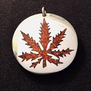May include: A silver pendant in the shape of a marijuana leaf with a red and orange sand-like material filling the leaf design.