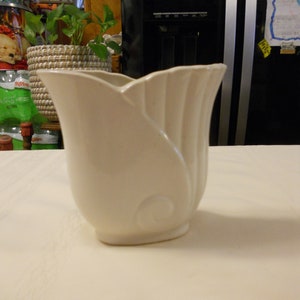 May include: A white ceramic vase with a unique, modern design. The vase has a wide, open mouth and a narrow base. It features a smooth, glossy finish.