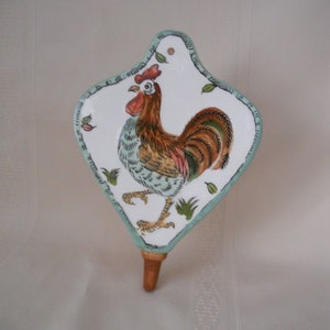 May include: A white ceramic rooster wall decor with green and brown accents. The rooster is depicted in a standing position with its wings spread out. The decor has a diamond shape with a wooden peg at the bottom.