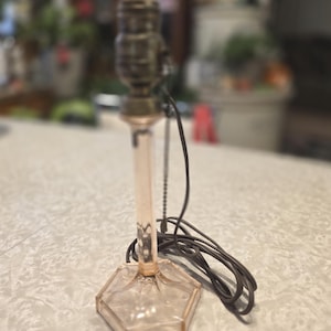 May include: A vintage pink glass table lamp with a hexagonal base and a slender stem. The lamp has a brass-colored socket and a black electrical cord. The lamp is approximately 12 inches tall.