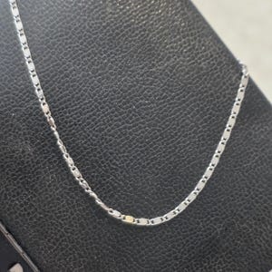 May include: A delicate silver necklace with a repeating rectangular link design. The chain is displayed against a dark background, highlighting its subtle shine. A simple and elegant piece of jewelry.