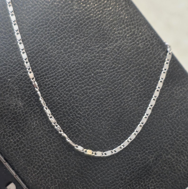 May include: A delicate silver necklace with a repeating rectangular link design. The chain is displayed against a dark background, highlighting its subtle shine. A simple and elegant piece of jewelry.