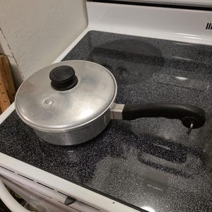 May include: A small, aluminium saucepan with a lid and a black handle. The pot is on a black stovetop. The lid has a black knob. The stovetop has a white oven with knobs and the word "Maytag" on it.
