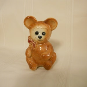 May include: A brown ceramic teddy bear figurine with a red bow. The bear has black eyes and a black nose.