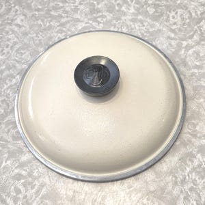 May include: Cream-colored pot lid with a silver rim and a dark gray handle. The handle has the word "Club" embossed on it. The lid is round and appears to be made of metal.