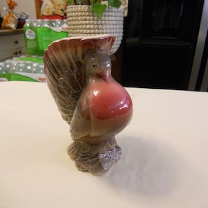 May include: A ceramic turkey figurine with brown and grey feathers, a red head, and a white base. The turkey is standing upright with its head tilted slightly to the side.