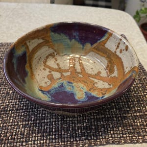May include: A handmade ceramic bowl with a wide rim. The bowl features a speckled off-white interior with brown, blue, and orange abstract designs. The exterior rim is a deep purple color. This unique bowl is perfect for serving food.