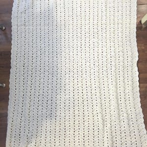 May include: Cream-colored crochet blanket with a scalloped edge. The blanket features a textured pattern of vertical lines and small openings throughout. The handmade blanket is laid out on a wooden surface.