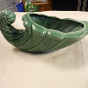 May include: A green ceramic planter shaped like a leaf with a curled handle. The planter has a smooth, glossy finish.