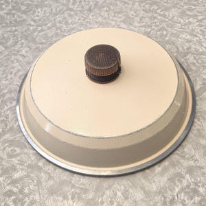 May include: Cream-coloured, round lid with a dark wooden knob. The lid has a silver rim and a slightly textured surface. The knob is made of dark wood with visible grain. The lid appears to be part of a cookware set.