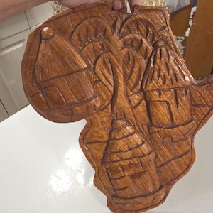 May include: A hand-carved wooden decorative piece in the shape of the African continent. The brown wood features detailed carvings of huts and palm trees, showcasing a rustic, natural aesthetic. The piece is held by a person.