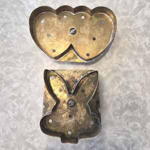 May include: Two vintage metal cookie cutters. One is a double heart shape, and the other is a bunny rabbit. Both cutters have a tarnished brass finish and small holes for easy release.