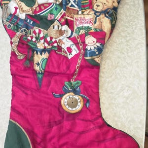 May include: A quilted Christmas stocking in deep pink with a dark green cuff and toe. The fabric features a festive print of Santa, a rocking horse, a teddy bear, and a book titled The Night Before Christmas. A gold pocket watch charm hangs.