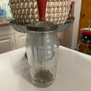 May include: A vintage glass jar with a red wooden top and a metal lid. The jar has a textured surface and is clear glass. The jar is used for storing and dispensing food.