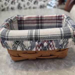 May include: A small, rectangular woven basket with a plaid fabric lining. The basket is made of light brown wood and features a fabric liner with a pattern of white, gray, and burgundy checks.