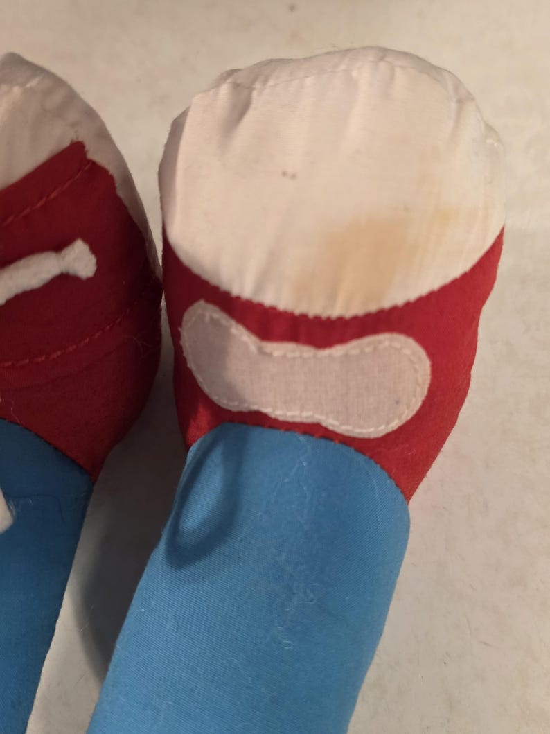 Peut inclure: Gros plan sur le pied et la jambe d'un jouet en peluche. Le pied est blanc avec une bordure rouge et un patch blanc. La jambe est bleue. Le jouet semble usag&eacute;, avec une certaine d&eacute;coloration.