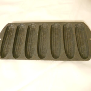 May include: A vintage cast iron cornbread pan with six individual moulds. The pan is dark grey and has a textured surface.