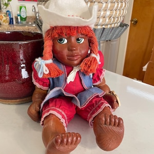 May include: A handmade doll with brown skin, red hair, and blue eyes. She is wearing a white hat, a red and blue dress, and a denim vest.