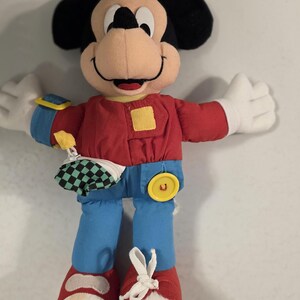 Peut inclure: Poup&eacute;e en peluche Mickey Mouse avec une chemise rouge, un pantalon bleu et des chaussures rouges. La poup&eacute;e a des oreilles noires, un visage bronz&eacute; et des gants blancs. Elle a un bouton jaune et une petite pochette &agrave; carreaux.