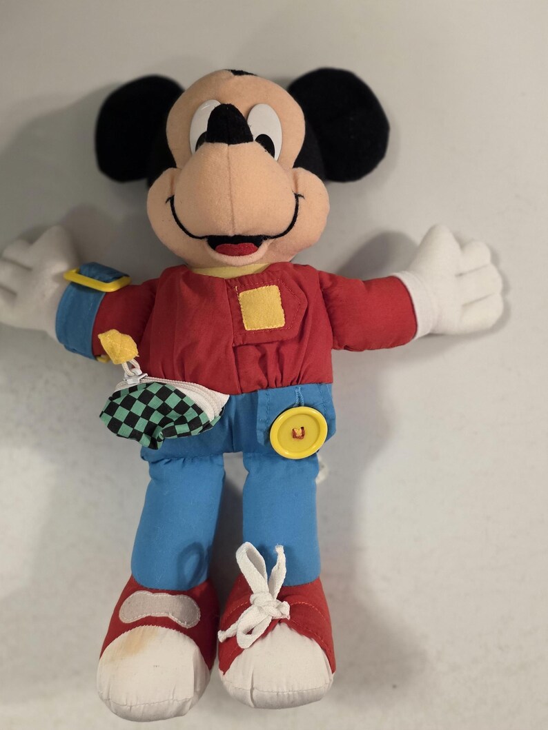 Peut inclure: Poup&eacute;e en peluche Mickey Mouse avec une chemise rouge, un pantalon bleu et des chaussures rouges. La poup&eacute;e a des oreilles noires, un visage bronz&eacute; et des gants blancs. Elle a un bouton jaune et une petite pochette &agrave; carreaux.