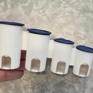 May include: A white plastic spice rack with four cylindrical containers. Each container has a dark blue lid and a clear, rectangular window at the bottom. The rack is held by a white horizontal bar.