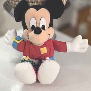Peut inclure: Une peluche Mickey Mouse avec des oreilles noires, une veste rouge, un pantalon bleu et des gants blancs. La poup&eacute;e a une expression souriante et est assise, les bras tendus. Un patch carr&eacute; jaune est sur la veste.
