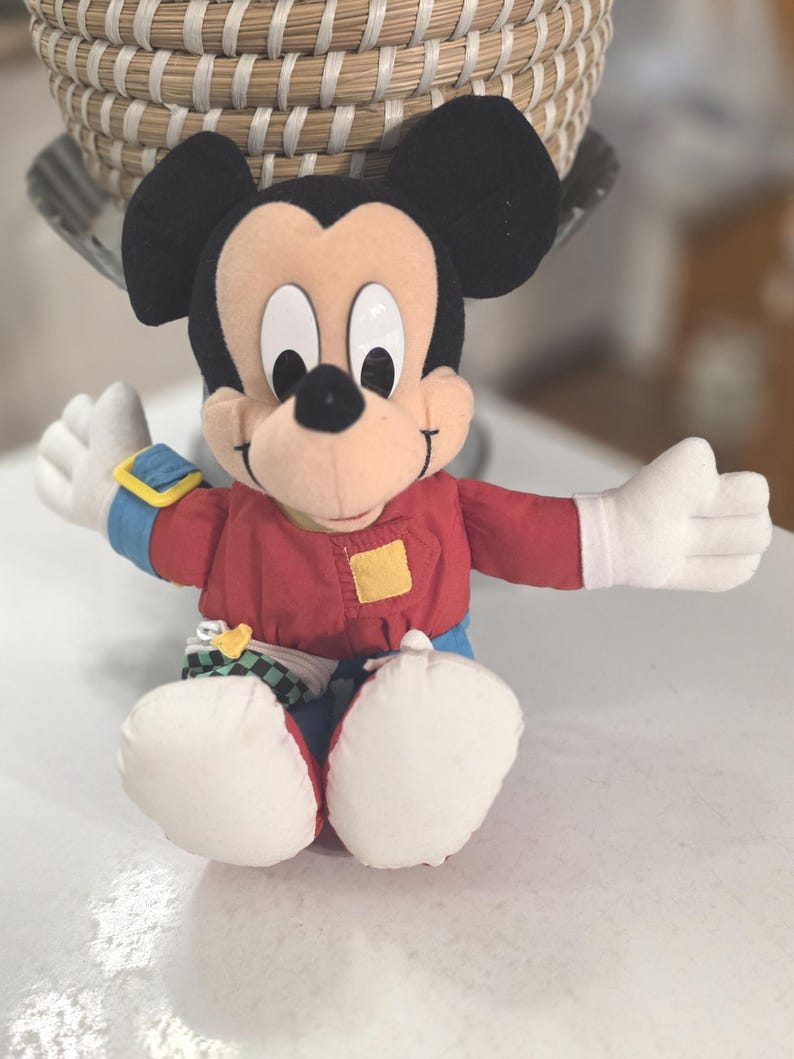 Peut inclure: Une peluche Mickey Mouse avec des oreilles noires, une veste rouge, un pantalon bleu et des gants blancs. La poup&eacute;e a une expression souriante et est assise, les bras tendus. Un patch carr&eacute; jaune est sur la veste.