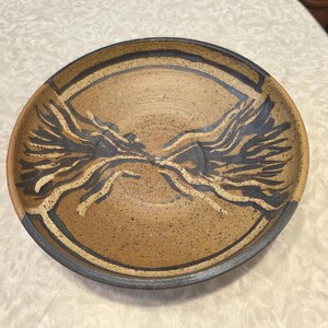 May include: A round, ceramic bowl with a speckled tan interior and a dark blue rim. The bowl features a stylized, abstract design in dark blue and cream colors, resembling a symmetrical, branching pattern. The bowl is suitable for serving food.