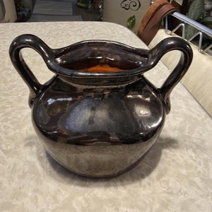 May include: A dark, metallic-glazed ceramic pot with two arched handles. The pot has a rounded body and a wide, wavy rim. The interior is a warm orange color, contrasting with the dark exterior. This decorative piece could be used as a vase or a decorative accent.