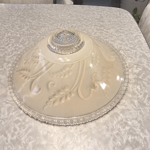 May include: A vintage, cream-colored glass lampshade with a decorative leaf and swirl pattern. The shade has a clear, textured rim and a clear glass center. The lampshade is round and appears to be in good condition.