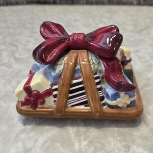 May include: A ceramic decorative piece shaped like a stack of wrapped gifts, topped with a large, glossy burgundy bow. The gifts are in various colors, including blue, white, and green, and are held together by brown wooden supports.