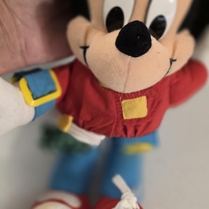 Peut inclure: Une peluche vintage de Mickey Mouse. La peluche porte une chemise rouge, un pantalon bleu et des chaussures rouges. Le jouet a un nez noir, de grands yeux noirs et une expression souriante. Le jouet est tenu par une personne.